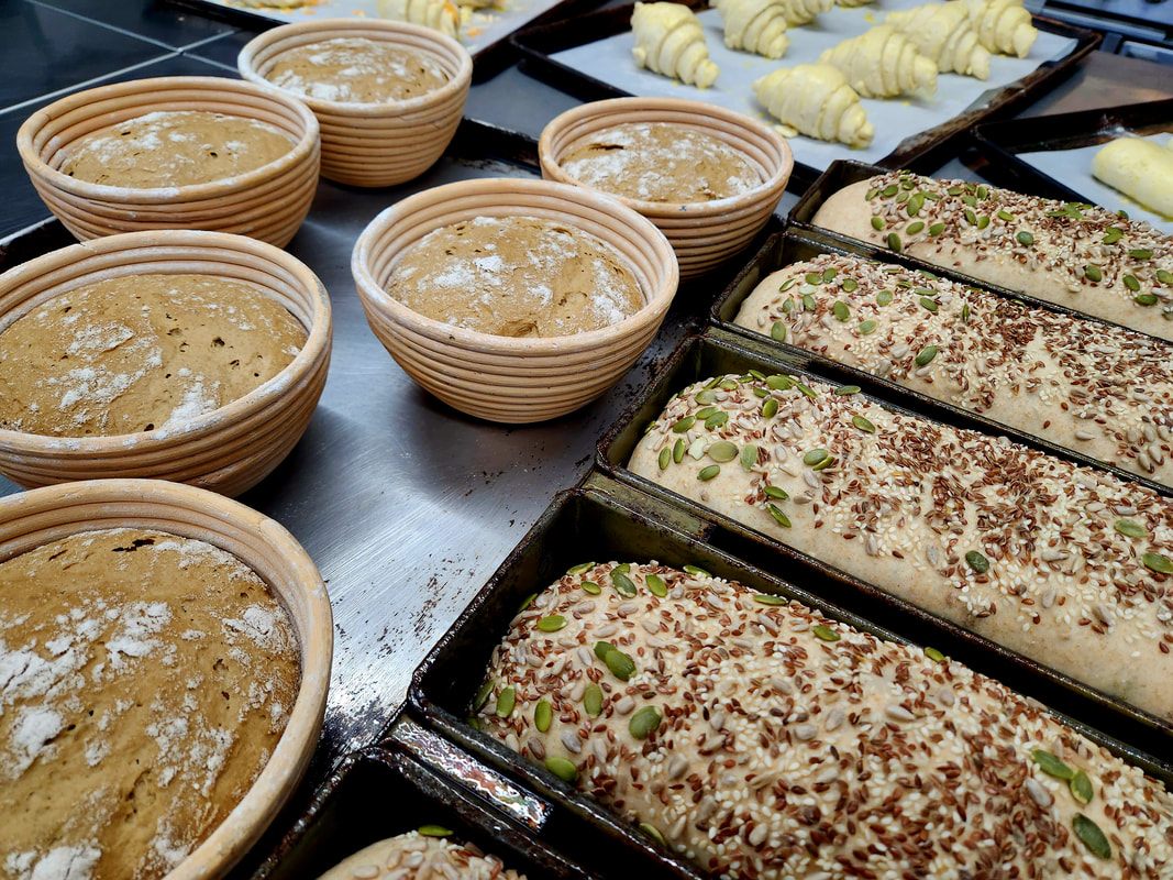 Seeded sourdough loaves in tins beside dough proofing in rattan baskets